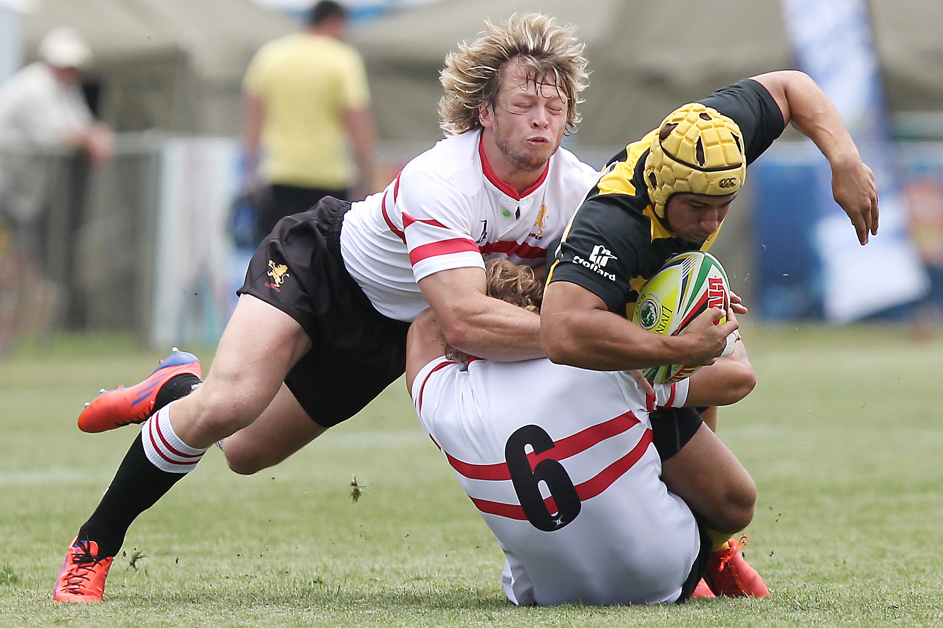 Two rugby players in white tackle an opposing player on the field who wears a protective yellow helmet.